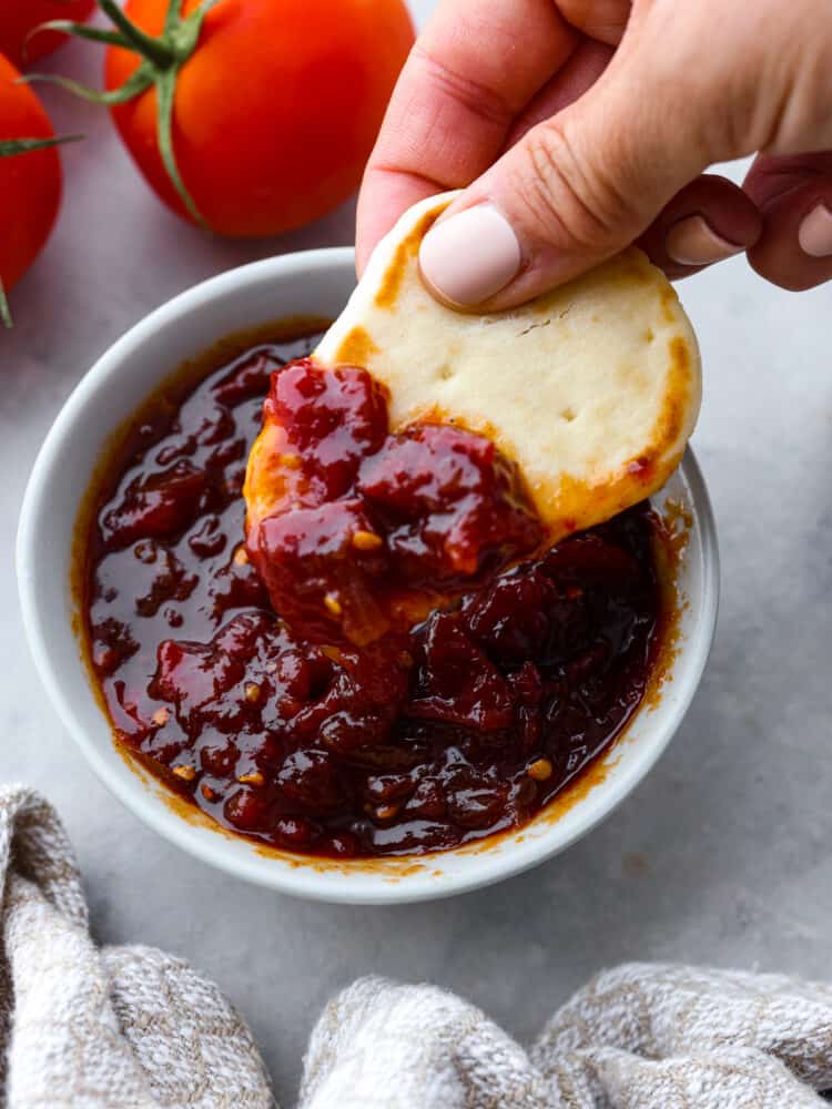 Tomato chutney is a sweet and savory condiment bursting with tomato flavor, delicious spices, vinegar, and brown sugar. Best known for being the perfect pairing for Indian cuisine, tomato chutney is a great addition to curries, naan bread, and charcuterie boards. Photo of a piece of naan bread being dipped into a bowl of tomato chutney. Tomatoes and a kitchen towel are styled next to the bowl of chutney.