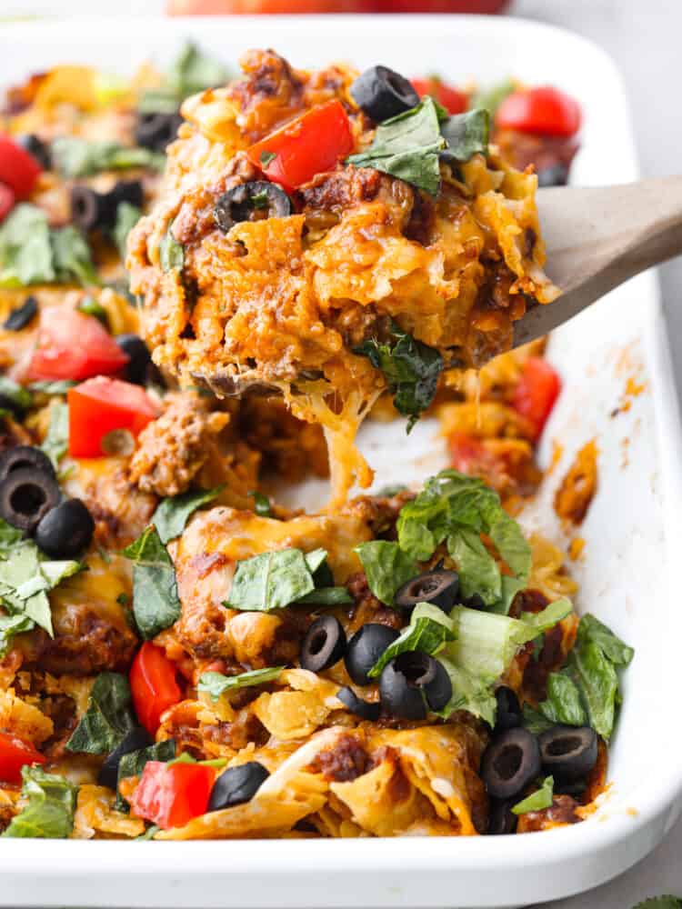 Easy taco casserole is beef, cheese, and tortilla chips layered up and loaded with rich flavor throughout. It is an easy-to-make, crowd-pleasing dinner that will be a hit with everyone! Taco casserole being scooped out with a wooden spoon.