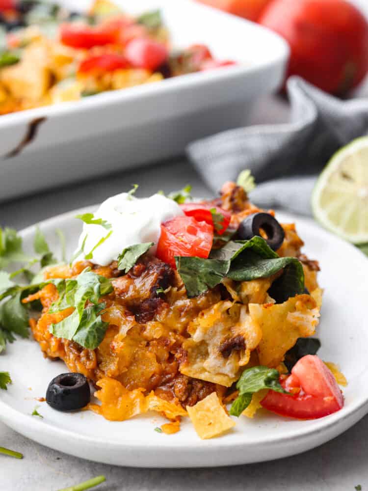 Easy taco casserole is beef, cheese, and tortilla chips layered up and loaded with rich flavor throughout. It is an easy-to-make, crowd-pleasing dinner that will be a hit with everyone! Some taco casserole on a plate loaded with veggies and sour cream.