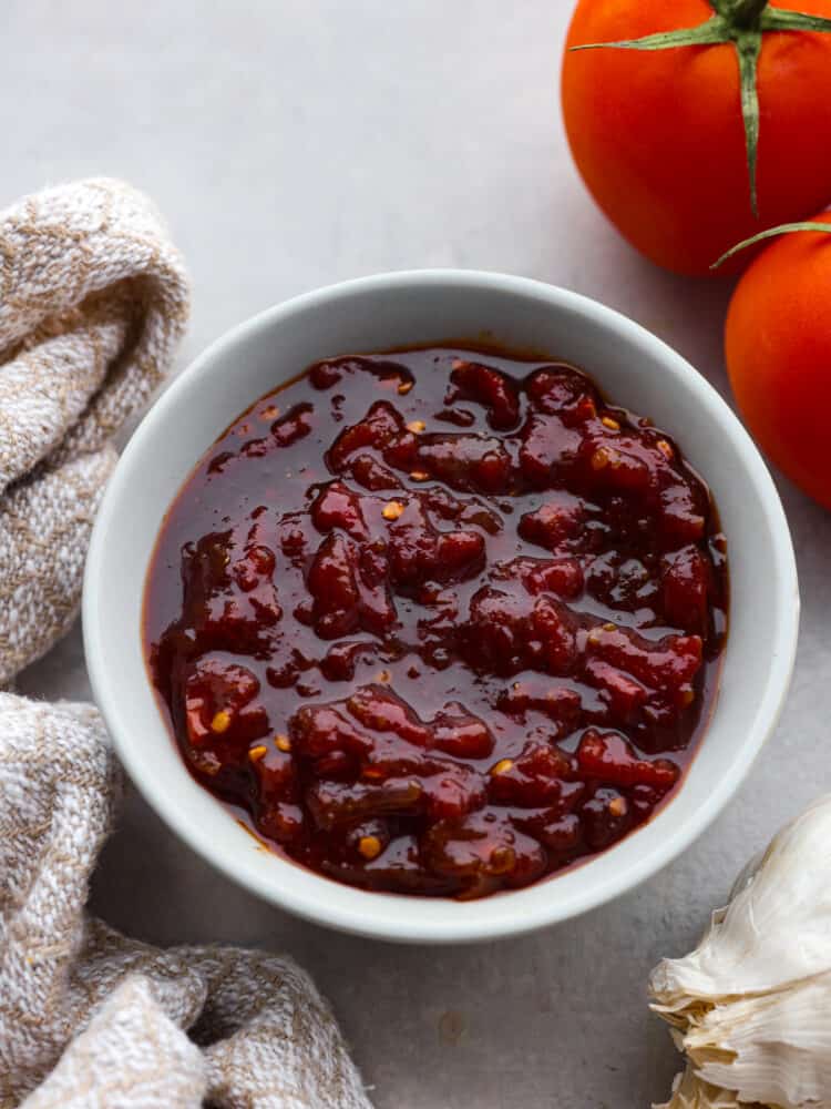 Tomato chutney is a sweet and savory condiment bursting with tomato flavor, delicious spices, vinegar, and brown sugar. Best known for being the perfect pairing for Indian cuisine, tomato chutney is a great addition to curries, naan bread, and charcuterie boards. Top view of tomato chutney in a small white bowl with tomatoes and kitchen towel next to the bowl.