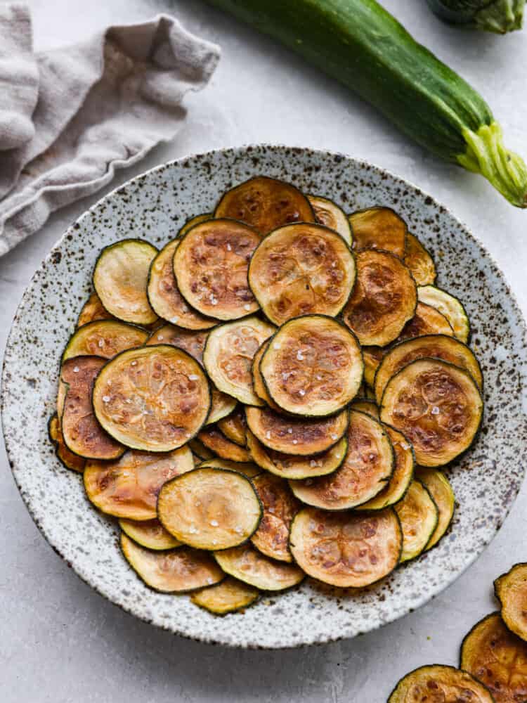 Need something to do with all of that fresh zucchini in your garden? I highly recommend you try this simple recipe for homemadeย Zucchini Chips! Simple to make and baked in the oven, these are a light, fun snack! A speckled plate with zucchini chips on it.