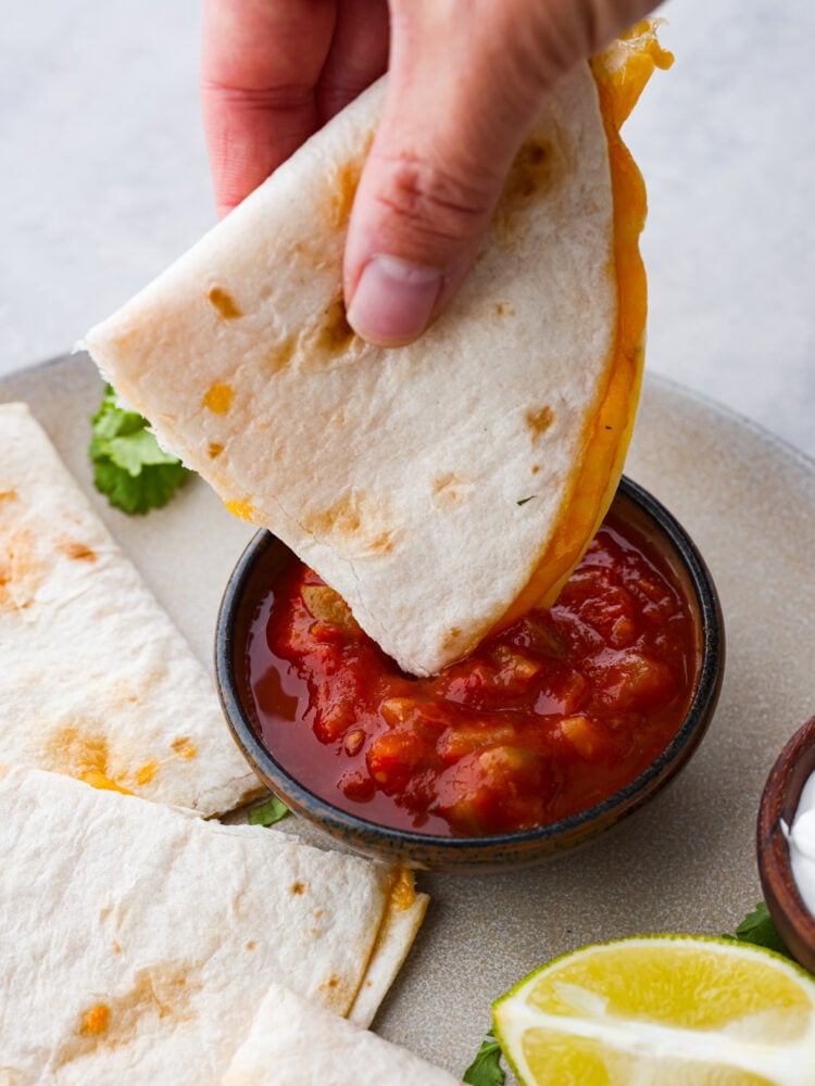 This air fryer quesadilla is the BEST and ONLY way to make a quesadilla. A perfectly crisp tortilla on the outside bursting with cheesy goodness on the inside, you will love how easy you can make a quesadilla in just minutes! Close up photo of a piece of quesadilla being dipped into a small bowl of salsa. The quesadilla and salsa are on a large gray plate. Cilantro and a lime wedge are garnished on the plate.
