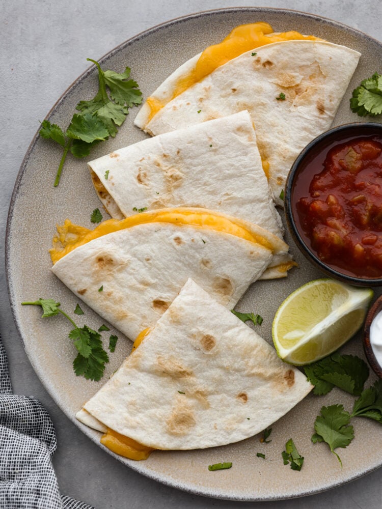 This air fryer quesadilla is the BEST and ONLY way to make a quesadilla. A perfectly crisp tortilla on the outside bursting with cheesy goodness on the inside, you will love how easy you can make a quesadilla in just minutes! Top view of an air fryer quesadilla cut into triangles on a large gray plate. A small bowl of salsa, cilantro leaves, and a lime wedge are also on the plate next to the quesadilla.
