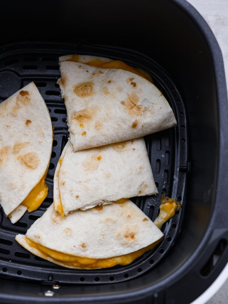 This air fryer quesadilla is the BEST and ONLY way to make a quesadilla. A perfectly crisp tortilla on the outside bursting with cheesy goodness on the inside, you will love how easy you can make a quesadilla in just minutes! Top view of air fryer quesadilla in a black air fryer basket. The quesadilla is cut into triangles with melted cheese oozing out.
