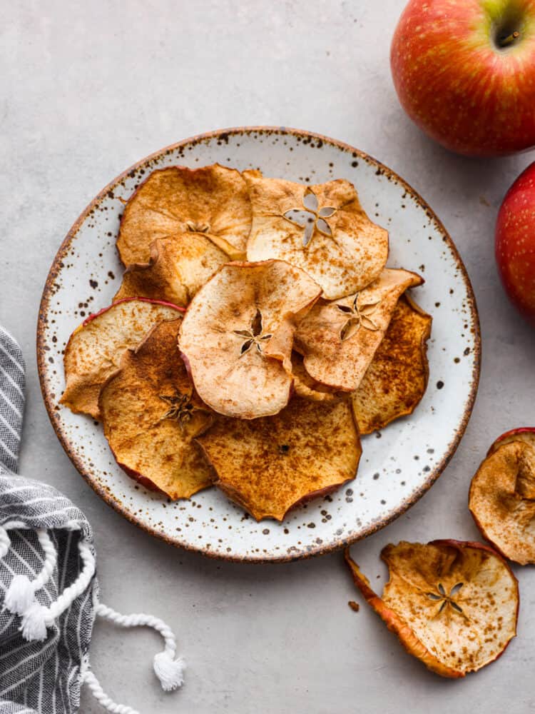 Air fryer apple chips are a healthy and delicious snack easily made in the air fryer. All you need is apples and spices and you can enjoy the sweet crisp of apple chips in just minutes! A pile of apple chips stacked on a small speckled plate. Red apples and a towel are styled by the plate.