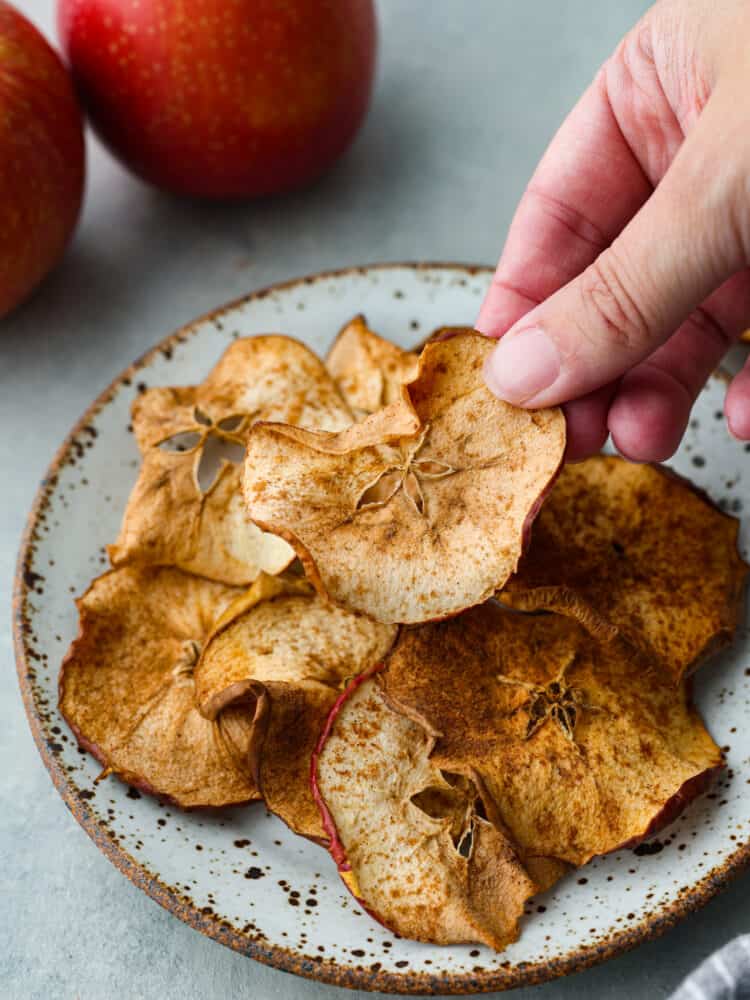 Air fryer apple chips are a healthy and delicious snack easily made in the air fryer. All you need is apples and spices and you can enjoy the sweet crisp of apple chips in just minutes! Close up photo of apple chips on a small plate. A hand is picking up an apple chip. Red apples are styled by the plate.