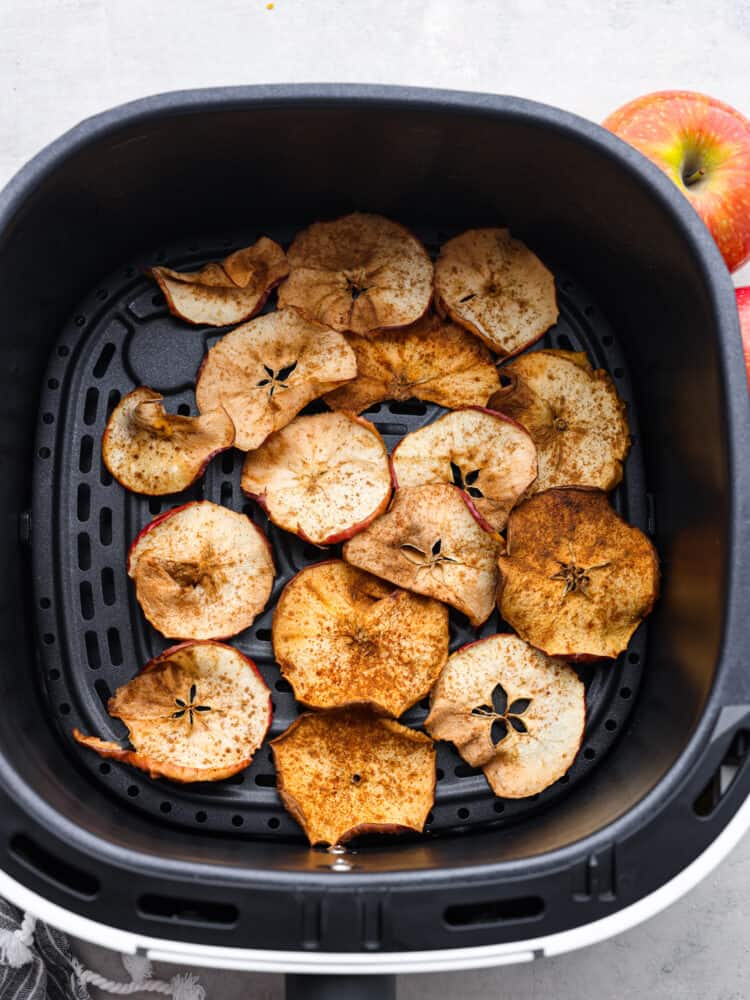 A photo of apple chips in the air fryer with apples next to the basket. Top view of spiced apple chips in the air fryer. Red apples are placed next the air fryer.