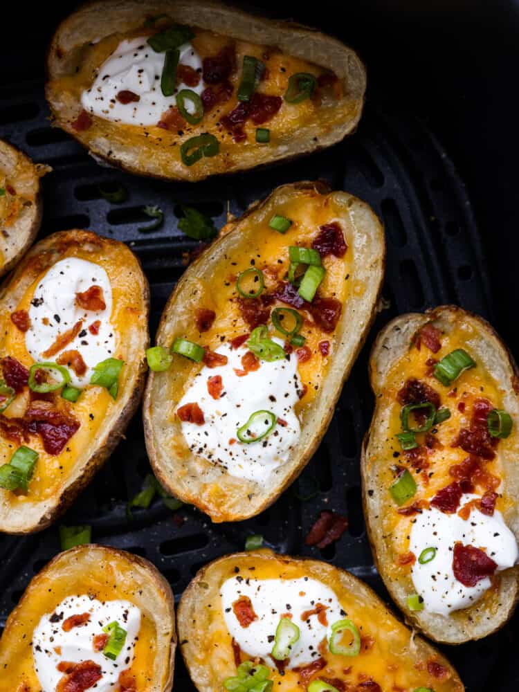 Air fryer potato skins are a quick and easy appetizer or side dish that everyone will love! Leftover baked potatoes turn into crispy and tender potato skins loaded with cheese, bacon crumbles, sour cream, and green onions. Close up view of air fryer potato skins in an air fryer garnished with sour cream, green onions, and bacon.