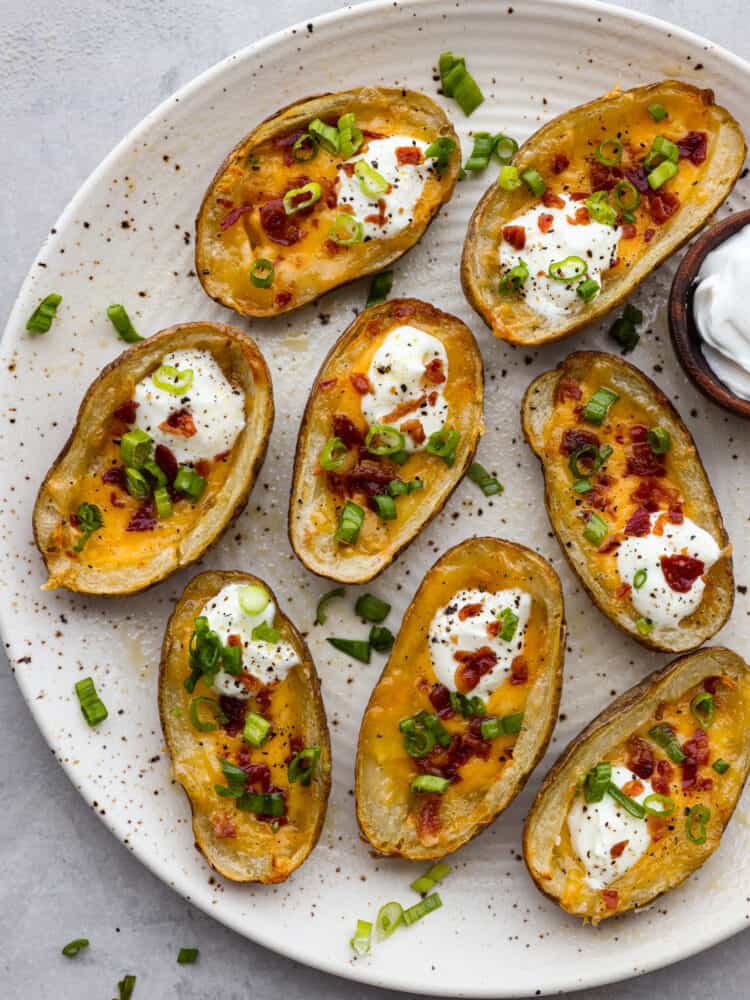 Air fryer potato skins are a quick and easy appetizer or side dish that everyone will love! Leftover baked potatoes turn into crispy and tender potato skins loaded with cheese, bacon crumbles, sour cream, and green onions. Top view of air fryer potato skins on a large white platter garnished with sour cream, green onions, and bacon. A small bowl of sour cream on the side.