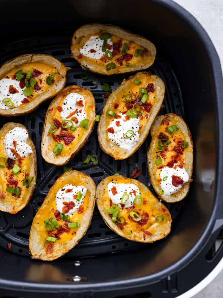 Air fryer potato skins are a quick and easy appetizer or side dish that everyone will love! Leftover baked potatoes turn into crispy and tender potato skins loaded with cheese, bacon crumbles, sour cream, and green onions. Top view of air fryer potato skins in an air fryer garnished with sour cream, green onions, and bacon.