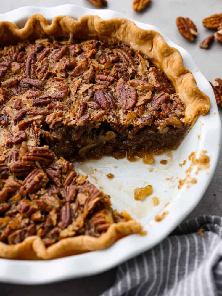 This gooey Bourbon Pecan Pie is a delicious twist on a classic! The alcohol will cook out while baking and you are left with rich flavor and a sweet filling that will leave your mouth watering. You won't want to make it any other way! Bourbon Pecan Pie with a slice taken out of it in a white pie dish with pecans in the background.