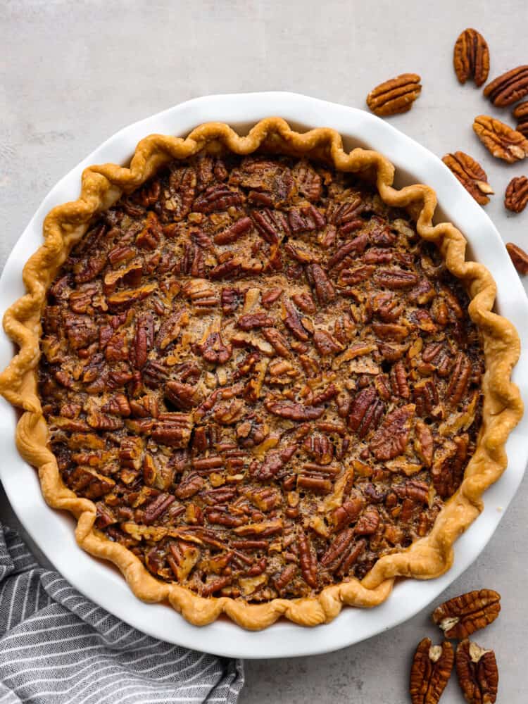 This gooey Bourbon Pecan Pie is a delicious twist on a classic! The alcohol will cook out while baking and you are left with rich flavor and a sweet filling that will leave your mouth watering. You won't want to make it any other way! A top view of a whole Bourbon Pecan Pie with a striped tea towel under neath and pecans sprinkled to the side.