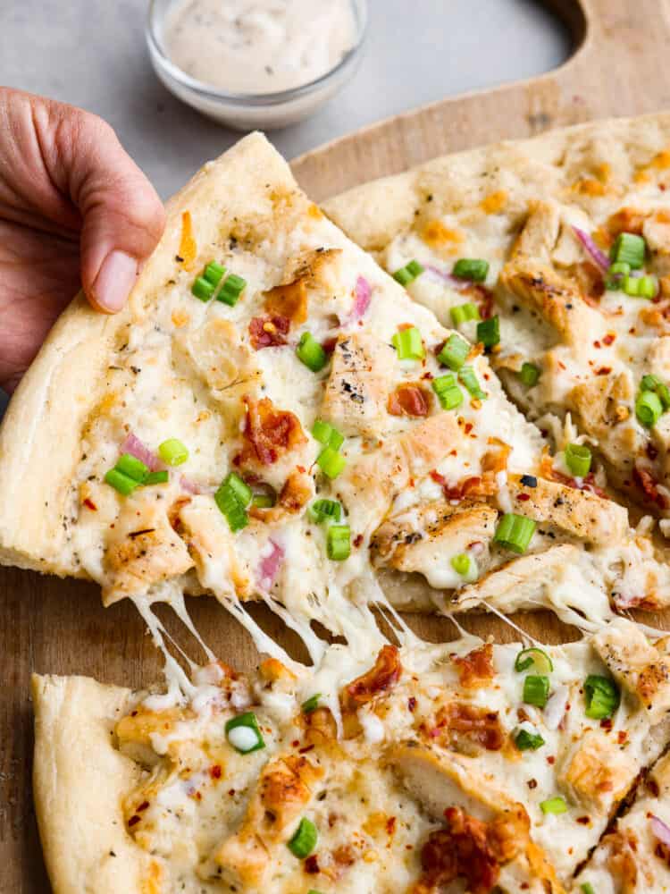 A total win for pizza night, this chicken alfredo pizza will be the best that you make. The homemade alfredo sauce is out of this worldย and the dough is quick and easy with only 20 minutes of rest time! Closeup of a slice of chicken alfredo pizza garnished with bacon and chopped green onions being picked up.