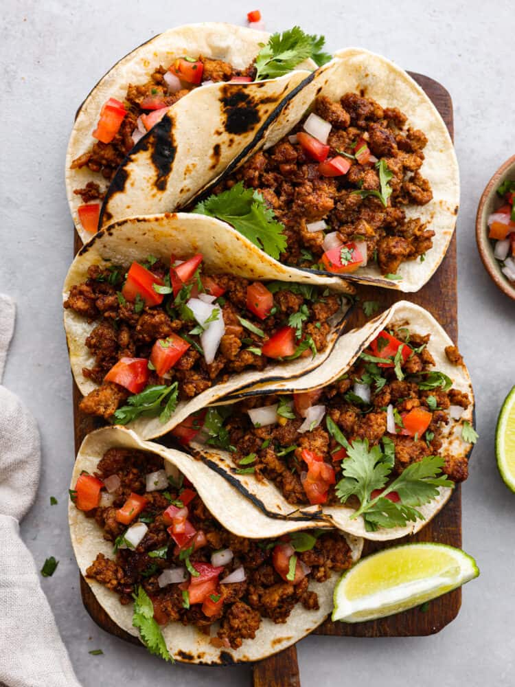 These Mexican Chorizo Tacos are the ultimate street food made right in your kitchen! Stuffed with mouthwatering Chorizo ground pork and topped with fresh herbs, this dish is sure to please a crowd. It only takes 15 minutes to make this meal that everyone will still be talking about tomorrow! Five Chorizo tacos sitting on a cutting board with tomatoes and onions and cilantro on top with limes to the side.