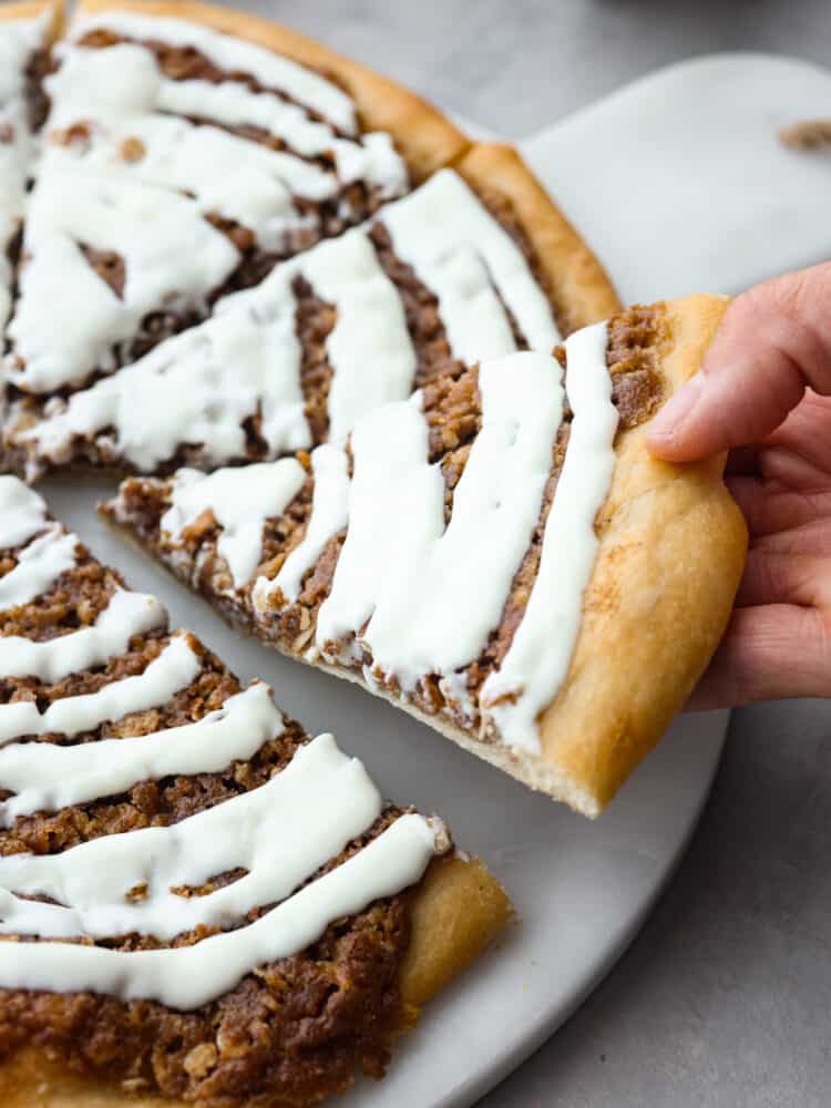 Inspired by Papa Murphy's cinnamon pizza, this dessert pizza has all of the goodness of a cinnamon roll! Buttery brown sugar cinnamon spread on top of quick and easy pizza dough and drizzled with a cream cheese glaze will make you go back for seconds! A slice of dessert pizza being taken off of the platter.