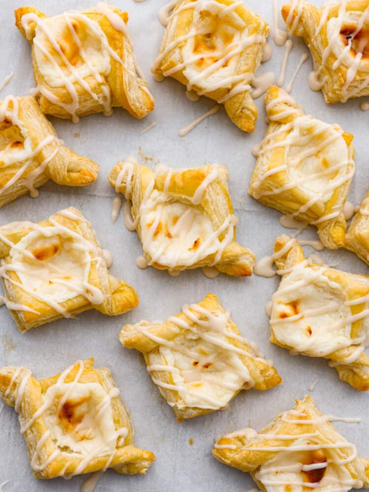 These cheese danishes are flaky, buttery, and full of tangy flavor. Topped with a sweet glaze and baked to perfection, this cheese danish recipe will make the perfect breakfast or dessert for any occasion. They were irresistibly good and gone way too fast! Closeup of baked cheese danishes laid out on parchment paper.