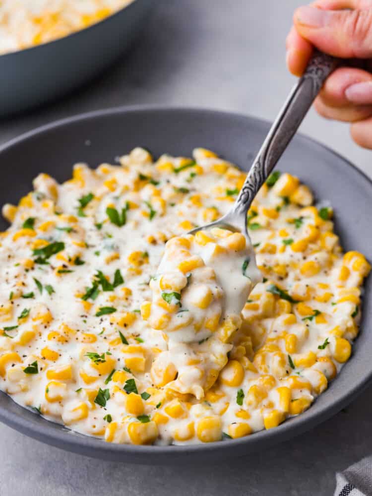 The ultimate side dish, this quick and easy creamed corn recipe is one you'll come back to again and again! It's buttery, creamy, and the perfect blend of savory and sweet! Closeup of creamed corn in a gray bowl being scooped up with a metal spoon.