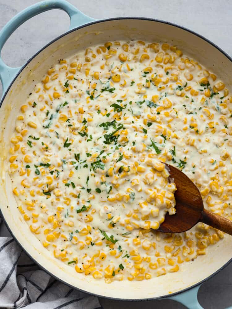 The ultimate side dish, this quick and easy creamed corn recipe is one you'll come back to again and again! It's buttery, creamy, and the perfect blend of savory and sweet! Top-down view of creamed corn, garnished with herbs in a white and blue skillet being stirred with a wooden spoon.