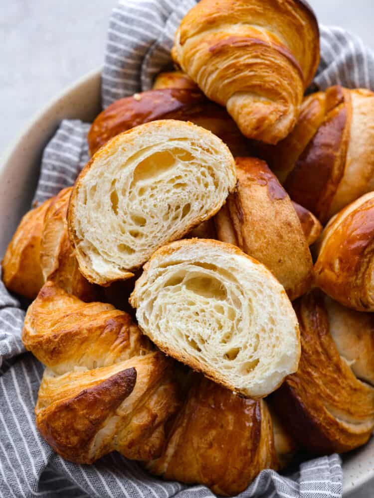 Close up view of croissants stacked on a gray striped kitchen towel and in a large cream colored bowl. One is cut and separated down the middle to show the baked inside layers.