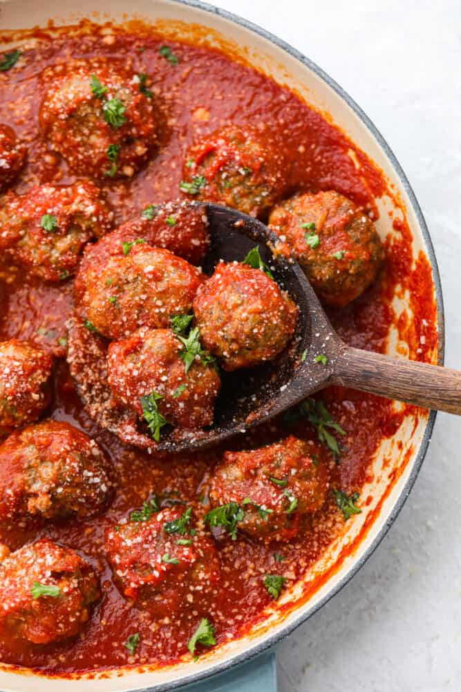 Whether you're making subs or spaghetti, these classic oven-baked meatballs are perfect for all of your favorite recipes. They're perfectly juicy and flavored with delicious seasonings and parmesan cheese! 3 meatballs covered in marinara sauce being scooped out of a skillet with a wooden spoon.
