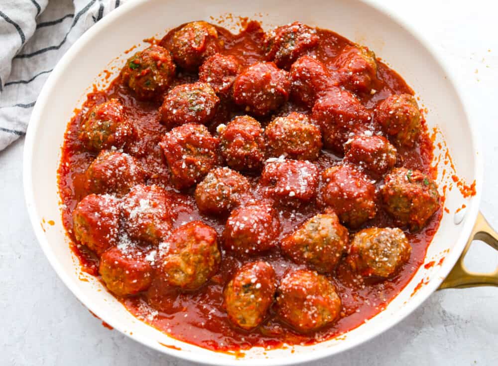 Whether you're making subs or spaghetti, these classic oven-baked meatballs are perfect for all of your favorite recipes. They're perfectly juicy and flavored with delicious seasonings and parmesan cheese! Cooked meatballs in marinara sauce, topped with grated parmesan in a white skillet.