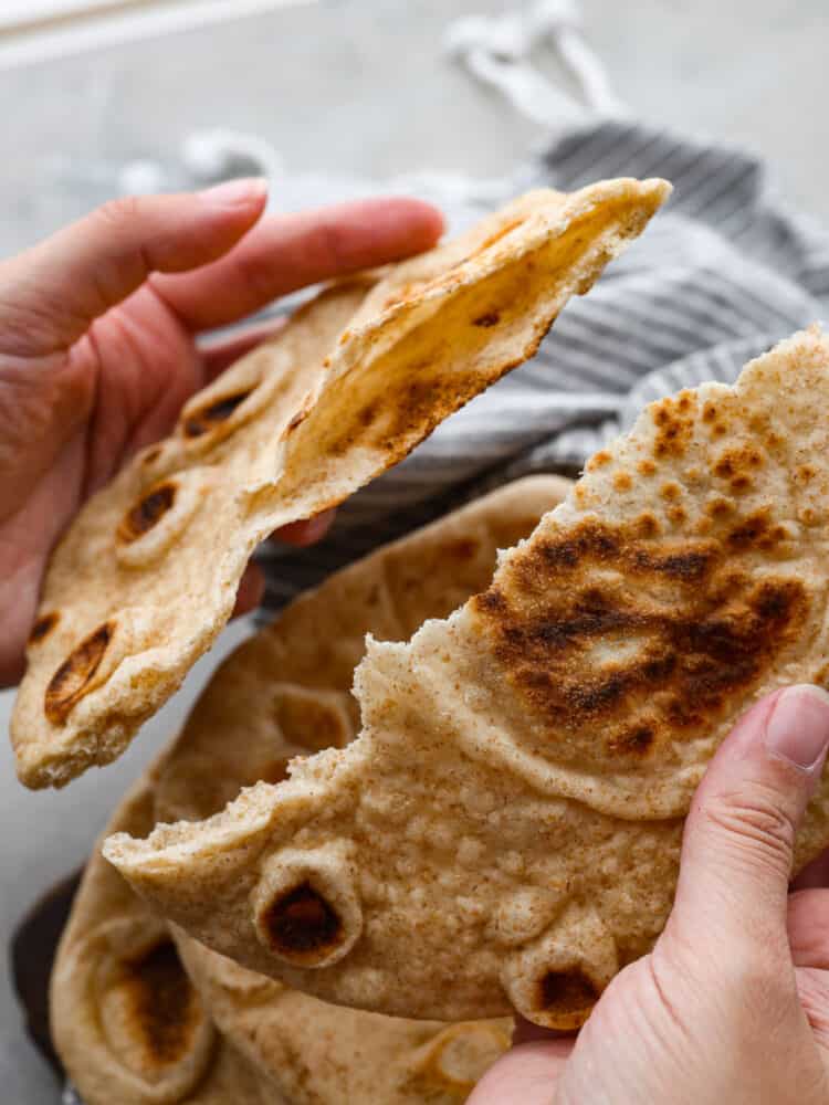 This homemade pita bread is soft, fluffy, and full of authentic flavor! Yes, you can buy pita bread at the store, but a freshly baked pita tastes so much better and will make your home smell absolutely delicious. Everyone will want to know what you are making! 2 hands breaking a piece of pita bread apart.