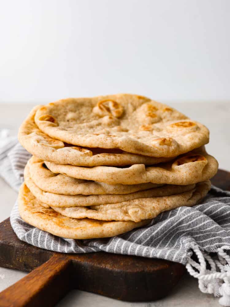 This homemade pita bread is soft, fluffy, and full of authentic flavor! Yes, you can buy pita bread at the store, but a freshly baked pita tastes so much better and will make your home smell absolutely delicious. Everyone will want to know what you are making! A stack of pita bread on a wooden cutting board with a blue striped tea towel.