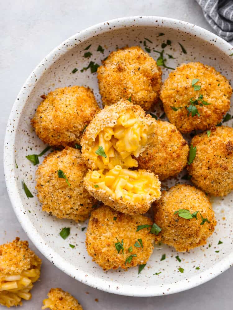These air fryer mac and cheese balls are crunchy, gooey, and full of delicious cheesy flavor! They are sure to be a hit with your kids or dinner guests, and you'll get this recipe requested again and again. A speckles, white bowl filled with Mac and cheese balls. One of them is broken open so that you can see the inside.
