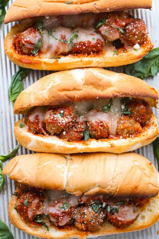 This easy meatball sub is always a favorite for both kids and adults. From the juicy meatballs smothered in marinara sauce to the toasted bun and melty cheese on top...it's the perfect sandwich for game day or any day! Three meatball sub sandwiches laying on a metal sheet pan. Melted cheese on top of the meatballs in a toasted hoagie roll. The subs are garnished with fresh basil leaves on the side.
