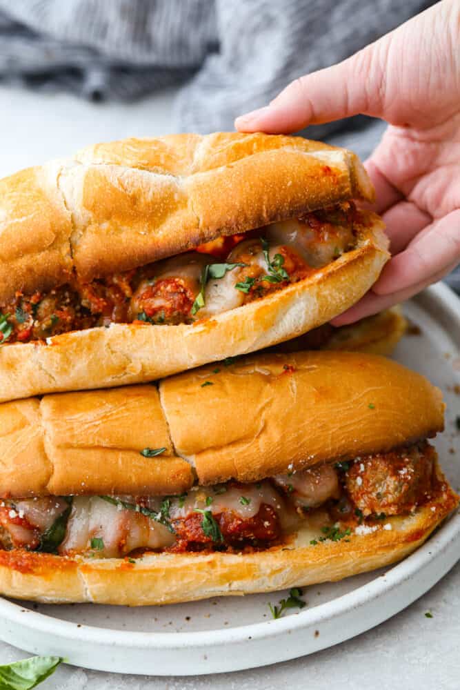 This easy meatball sub is always a favorite for both kids and adults. From the juicy meatballs smothered in marinara sauce to the toasted bun and melty cheese on top...it's the perfect sandwich for game day or any day! Close up view of a stack of meatball subs. A hand is reaching in and lifting up one of the subs on top.