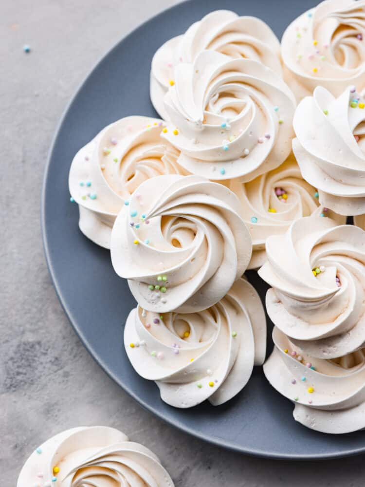 Meringue cookies are crispy on the outside and chewy on the inside. The whipped egg white base just melts in your mouth and tastes absolutely divine. They are a beautiful dessert for any occasion! Meringue cookies sitting on a blue plate with sprinkles on top.