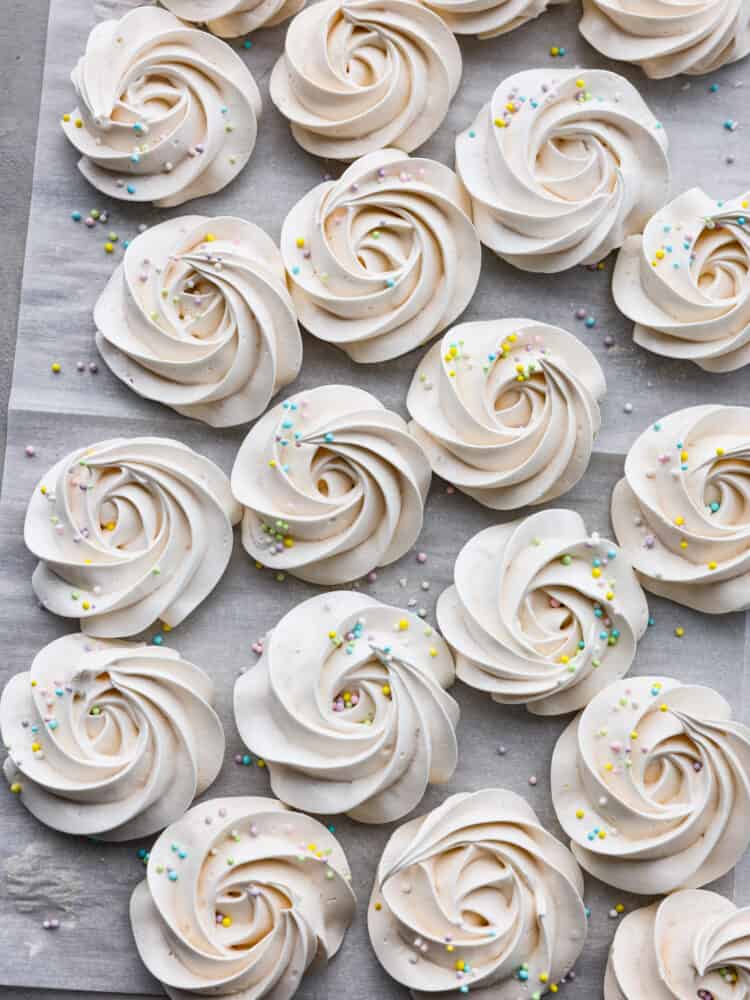 Meringue cookies are crispy on the outside and chewy on the inside. The whipped egg white base just melts in your mouth and tastes absolutely divine. They are a beautiful dessert for any occasion! A zoomed out shot of the cookies on parchment paper with sprinkles.