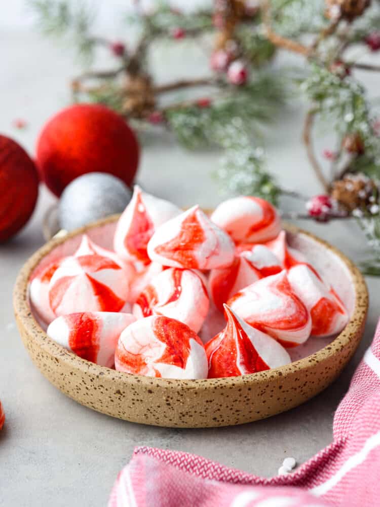 Peppermint Meringue Kisses are crunchy on the outside with a soft and chewy middle, and a peppermint flavor that is sweet and refreshing.ย They are the cutest little candies and are perfect for gifting this holiday season! Peppermint meringue kisses in a candy dish with greenery and christmas ornaments in the background.