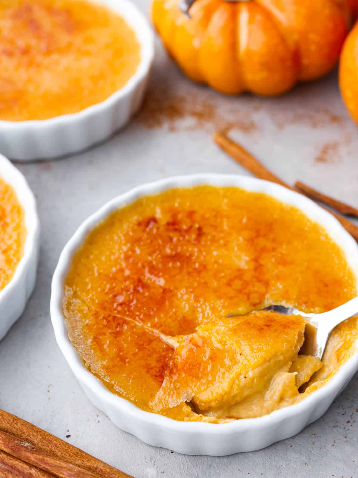 This creamy pumpkin crème brûlée adds a seasonal twist on a classic dessert. This delicious fall version of crème brûlée has pumpkin custard spiced to perfection and topped with a caramelized sugar coating. Close side view of a spoon in a ramekin of pumpkin crème brûlée.The caramelized top is cracked into with the spoon. A pumpkin and cinnamon sticks are next to the ramekins.