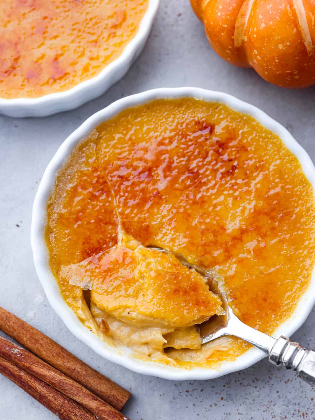 Close top view of a spoon in a ramekin of pumpkin crème brûlée. A pumpkin and cinnamon sticks are next to the ramekins.