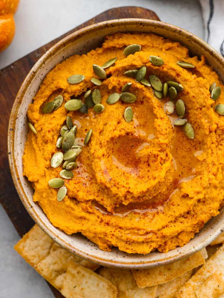 This spiced pumpkin hummus is the most delicious addition to all of your fall parties! Chickpeas, garlic, tahini, tasty seasonings, and rich pumpkin combine to create a flavorful fall appetizer you won't be able to get enough of. Top-down view of pumpkin hummus in a white bowl, garnished with pumpkin seeds.