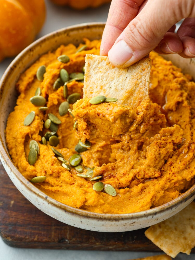 This spiced pumpkin hummus is the most delicious addition to all of your fall parties! Chickpeas, garlic, tahini, tasty seasonings, and rich pumpkin combine to create a flavorful fall appetizer you won't be able to get enough of. Closeup of a pita chip being dipped into a bowl of pumpkin hummus.