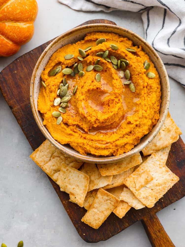 This spiced pumpkin hummus is the most delicious addition to all of your fall parties! Chickpeas, garlic, tahini, tasty seasonings, and rich pumpkin combine to create a flavorful fall appetizer you won't be able to get enough of. Pumpkin hummus served on a wooden tray with pita chips.