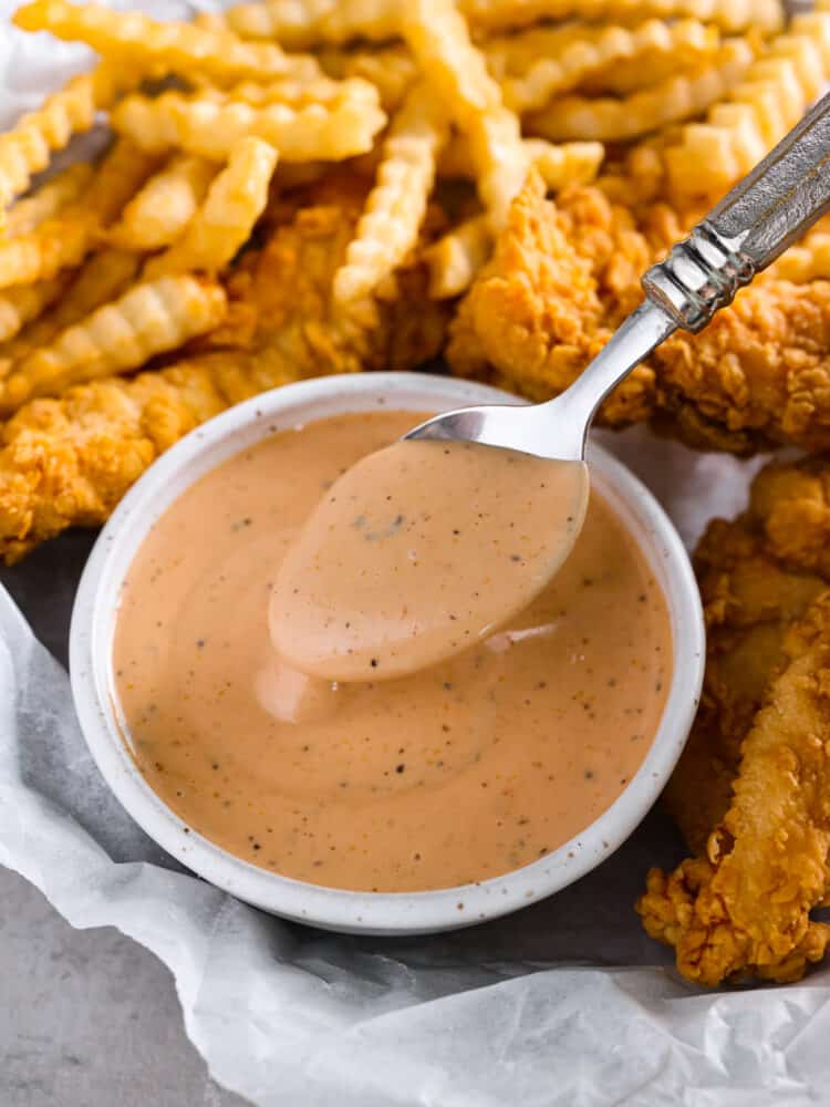 The popular cane's sauce from Raising Cane's restaurant can be made at home with this identical copycat version. This creamy and tangy dipping sauce is packed with savory smoky flavors perfect for chicken, fries, or as a burger spread. Close up view of sauce in a small white bowl on top of parchment paper. A spoon is lifting up sauce. French fries and chicken stripes are scattered around the bowl.