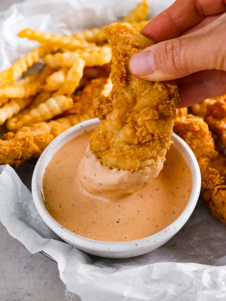 The popular cane's sauce from Raising Cane's restaurant can be made at home with this identical copycat version. This creamy and tangy dipping sauce is packed with savory smoky flavors perfect for chicken, fries, or as a burger spread. Close up view of a chicken finger being dipped into the bowl of sauce. French fries and chicken stripes are scattered around the bowl.