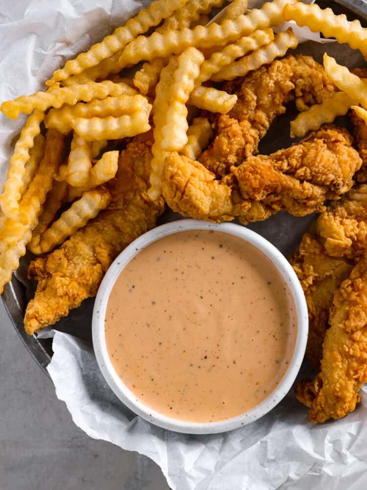 The popular cane's sauce from Raising Cane's restaurant can be made at home with this identical copycat version. This creamy and tangy dipping sauce is packed with savory smoky flavors perfect for chicken, fries, or as a burger spread. A top view of the sauce in a small white bowl. The bowl is on a round tin plate with parchment paper. Chicken fingers and crinkle cut fries are surrounding the bowl.