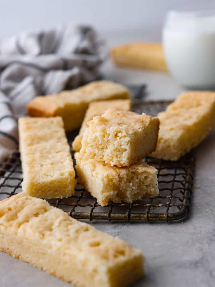 Traditional Scottish shortbread is buttery, crispy, and lightly sweetened to perfection. Simply made with butter, flour, sugar, and salt, this classic treat can easily be made for the holidays or any time of year. Close up view of Scottish shortbread stacked on a cooling rack. One cookie is broken into two pieces. A kitchen towel and glass of milk are in the distance.