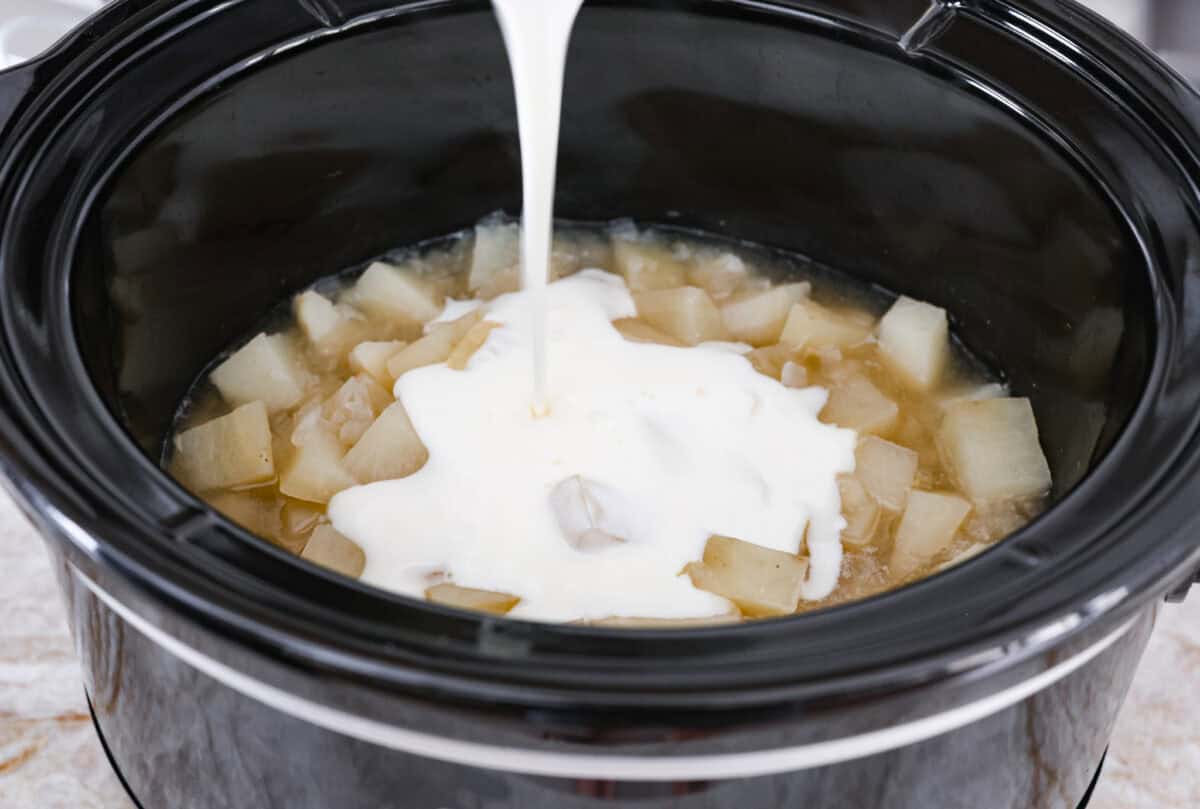 Loaded baked potato soup has a smooth, creamy base, soft potatoes, topped with cheese, bacon, green onion, and whatever else you like. It is hands down the best potato soup I have ever had. It is creamy and delicious, with all the goodness of a baked potato, made right in your slow cooker! Overhead shot of cream and roux mixture being poured over the onion and potatoes in the slow cooker.