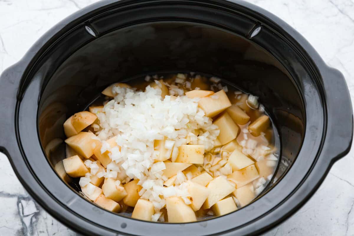 Loaded baked potato soup has a smooth, creamy base, soft potatoes, topped with cheese, bacon, green onion, and whatever else you like. It is hands down the best potato soup I have ever had. It is creamy and delicious, with all the goodness of a baked potato, made right in your slow cooker! Overhead shot of potatoes, onions and chicken broth in slow cooker.