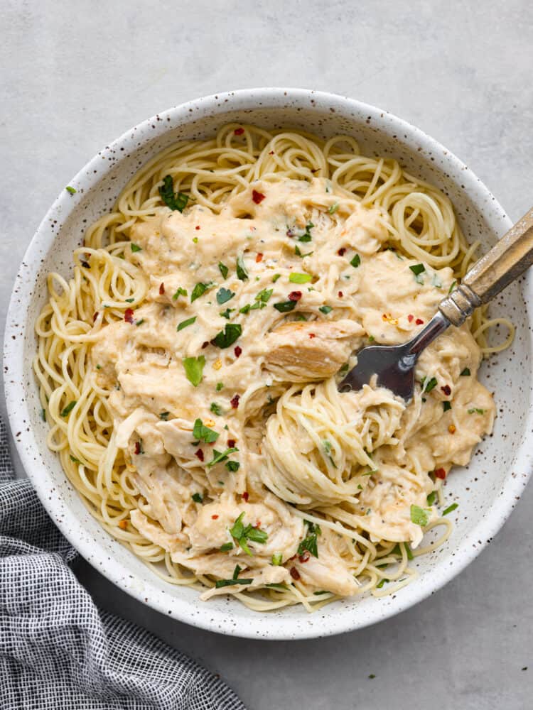 Slow cooker angel chicken is a quick and easy weeknight meal that your family will request all the time! Tender chicken cooked in a creamy Italian-flavored sauce and served on a bed of angel hair pasta. Top view of angel chicken in a white speckled bowl. Angel hair pasta is wrapped around a fork that is placed in the bowl. A gray kitchen towel is next to the bowl.