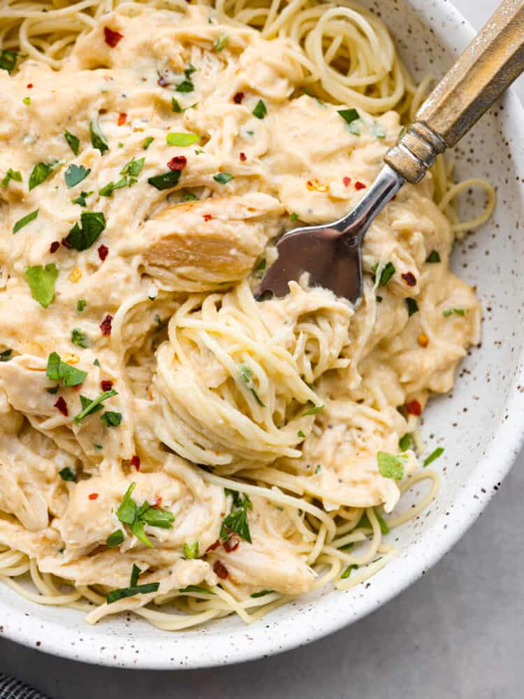 Slow cooker angel chicken is a quick and easy weeknight meal that your family will request all the time! Tender chicken cooked in a creamy Italian-flavored sauce and served on a bed of angel hair pasta. Close up photo of slow cooker angel pasta plated in a white speckled bowl. A fork is in the bowl with parsley and red pepper flakes garnished on top.