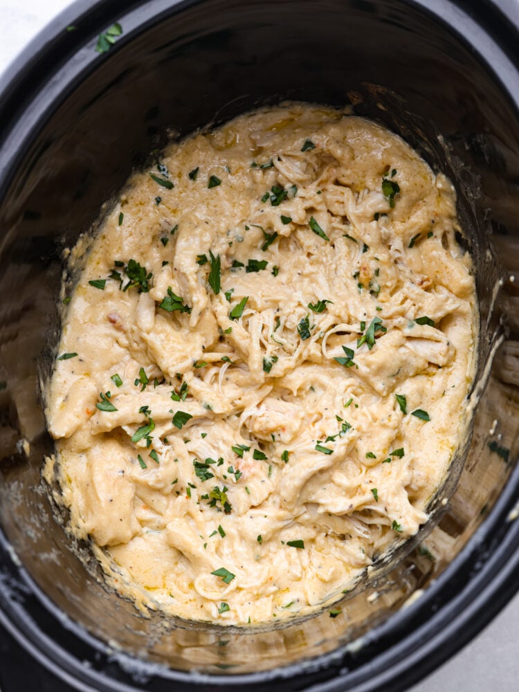Slow cooker angel chicken is a quick and easy weeknight meal that your family will request all the time! Tender chicken cooked in a creamy Italian-flavored sauce and served on a bed of angel hair pasta. Close up top view of cooked slow cooker angel chicken in a black crockpot. Fresh parsley is garnished on top.