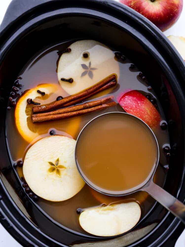 This easy crockpot apple cider is the must-have drink for this fall and winter! It has all the right spices and flavors and will warm you right up on those cold winter days! It's perfect for any holiday party or gathering! A crockpot filled with apple cider , a measuring cup, apple slices and cloves.