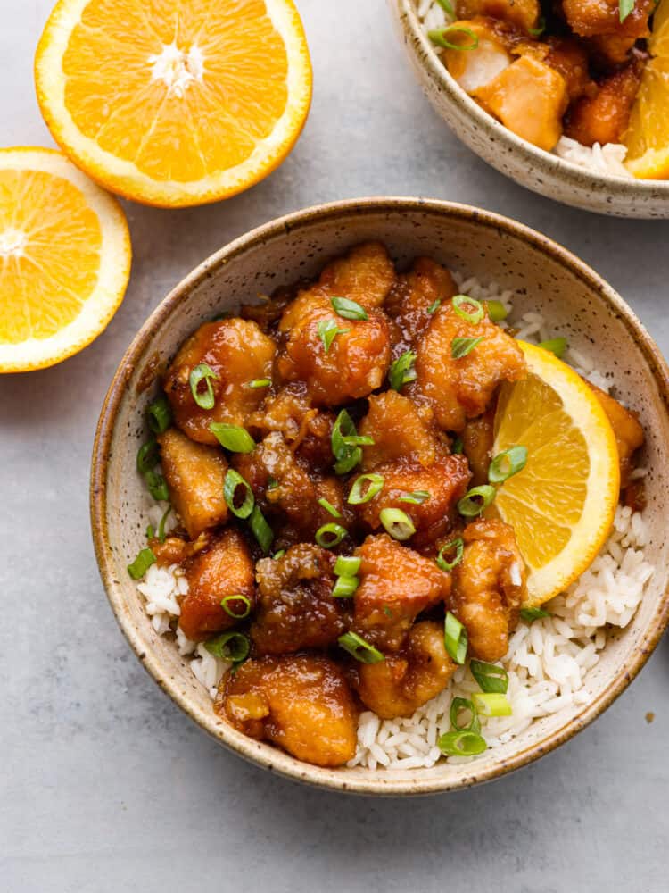 This delicious and tangy orange chicken is made right in your slow cooker! This is better than takeout! This tender chicken is coated in a irresistible sauce that will keep your family asking for this at least once a week! The top view of a bowl of orange chicken on rice with some orange slices.