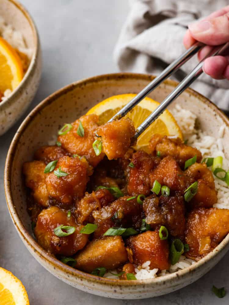 This delicious and tangy orange chicken is made right in your slow cooker! This is better than takeout! This tender chicken is coated in a irresistible sauce that will keep your family asking for this at least once a week! Orange chicken in a bowl with some silver chopsticks taking out a piece.